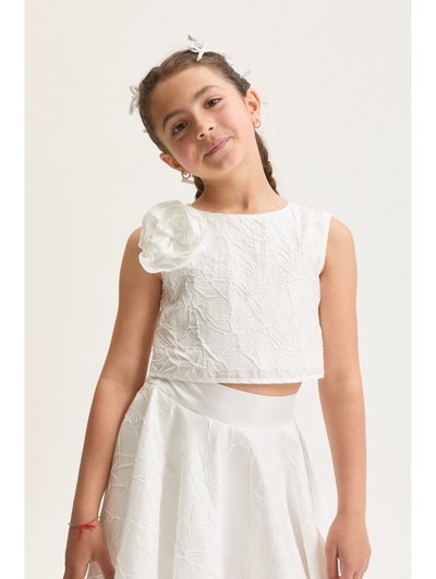 Ung pige med flettet hår bundet med hvide bånd, iført en hvid ærmeløs kjole med en stor stofblomst på skulderen.