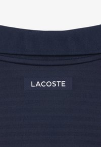 Tela texturizada azul marino con una etiqueta rectangular negra que muestra "LACOSTE" en letras mayúsculas blancas debajo de un cuello doblado.
