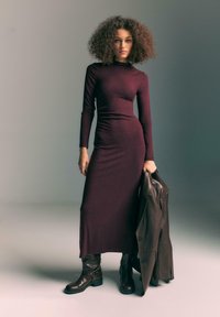 Burgundské šaty s dlhým rukávom a priliehavým strihom, midi dĺžka, jemná textúra. Modelka drží hnedú koženú bundu a má na sebe hnedé čižmy po kolená.