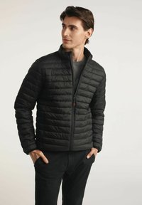 Czarna puchowa kurtka z poziomym pikowaniem, wysokim kołnierzem i zapięciem na zamek. Nosi ją osoba w szarej koszuli, do której ma czarne spodnie.