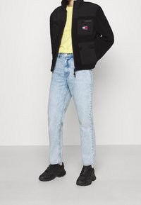 Czarna polarowa kurtka na żółtej koszulce, jasnoniebieskie dżinsy z wysokim stanem i czarne sportowe buty. Charakterystyczne naszywki i zamki w kurtce.