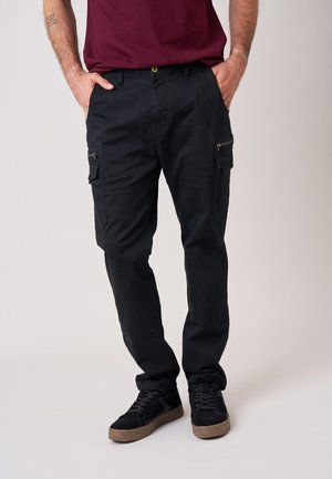 Man draagt zwarte cargobroek, zwarte sneakers met bruine zolen en een bordeauxrode t-shirt, staat met handen in de zakken tegen een effen achtergrond.