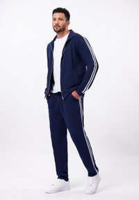 Survêtement bleu marine avec des rayures blanches, comprenant une veste zippée et un pantalon à taille élastique. Tissu texturé, associé à des baskets blanches.