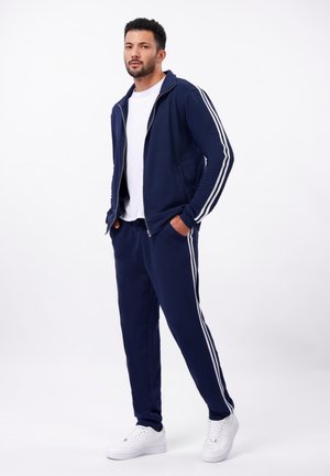 Navy tracksuit with white stripes, featuring a zip-up jacket and elastic waist pants. Textured fabric, paired with white sneakers.