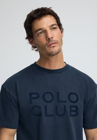 Marinblå oversized t-shirt i bomull, med stor text "POLO CLUB" i en något mörkare nyans. Rund halsringning och korta ärmar.