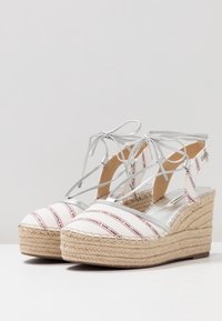 Białe koturny espadryle z czerwonymi paskami, srebrnymi wiązaniami wokół kostki oraz plecioną platformą z juty. Gładkie płótno z delikatnymi metalicznymi akcentami.