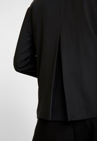 Veste noire à texture lisse, présentant un pli distinctif à l'arrière et une coupe courte, idéale pour superposer.