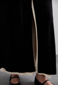 Lang sort kjole med en kontrasterende cremefarvet sidepanel og en slids, præget af glat stof og en enkel kant. Sorte sko med remdetaljer.