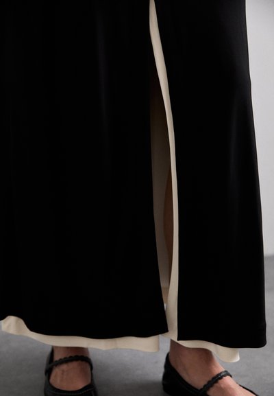 Long black dress with a contrasting cream side panel and a slit, featuring smooth fabric and a plain hem. Black shoes with strap accents.