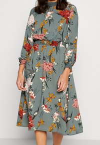 Abito floreale in tessuto verde, con maniche lunghe, vita arricciata e fiori di diversi colori tra cui accenti rossi, rosa e arancioni.