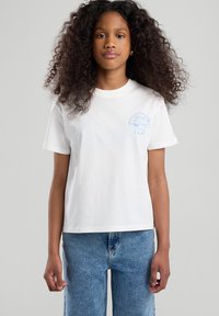 Valkoinen puuvillapaita, jossa on pyöreä kaula-aukko ja lyhyet hihat. Vasemmassa rinnassa on vaaleansininen graafinen kuvio. Yhdistetty sinisiin farkkushortseihin.