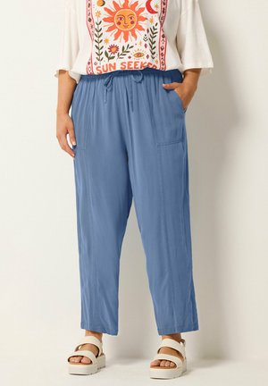 Kobieta nosząca jasnoniebieskie spodnie ze sznurkiem i dużymi przednimi kieszeniami, dobrane do białych sandałów oraz białego topu z grafiką o tematyce słonecznej.
