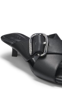 Μαύρο δερμάτινο mule με τακούνι, φαρδιές λουρίδες που διασταυρώνονται και μεγάλο ασημένιο αγκράφα σε λευκό φόντο.