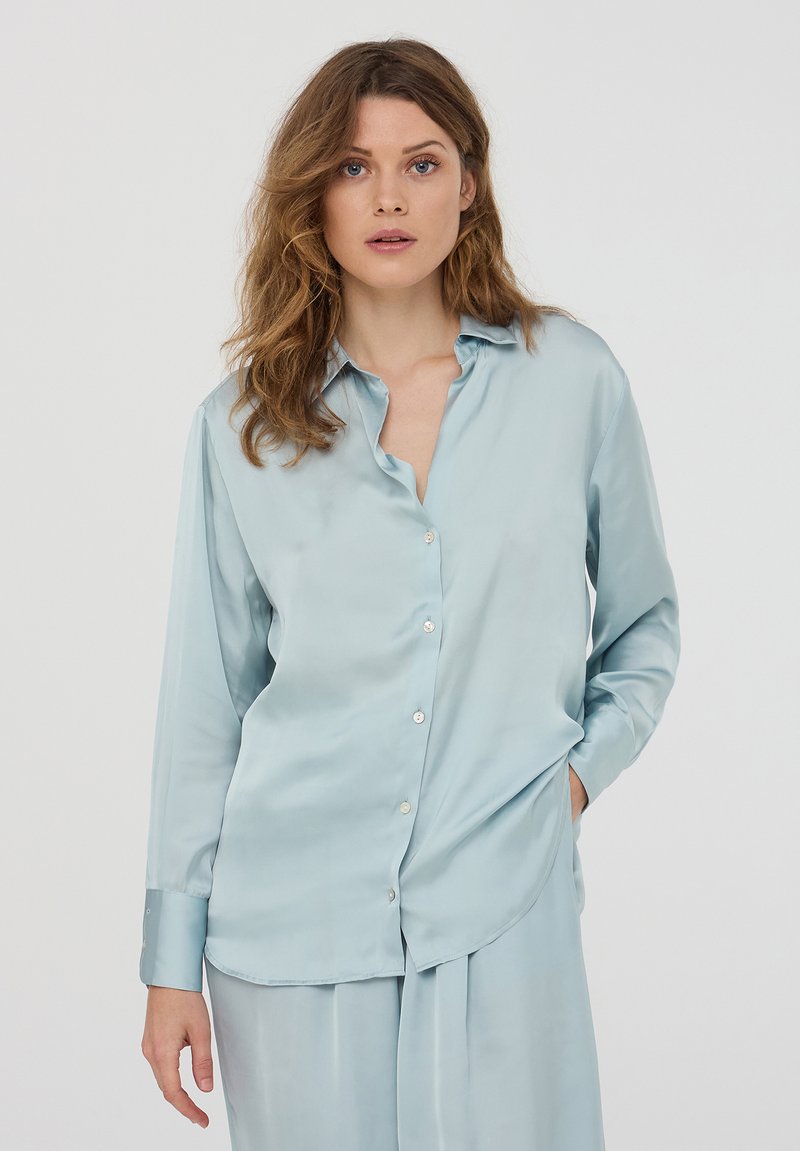 Camicia azzurra in satin con bottoni, maniche lunghe, colletto a punta e vestibilità comoda. Presenta una tasca frontale e bottoni lucidi.