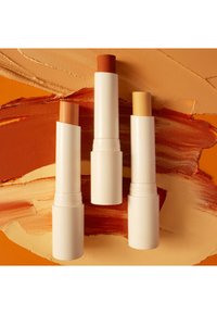 Tre prodotti per le labbra cilindrici in tubi bianchi, che mostrano tonalità arancioni, pesca e giallo chiaro con una texture liscia su uno sfondo arancione.