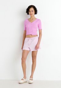 Top de punto corto y de manga corta en rosa claro, con botones en la parte delantera y bordes en forma de onda, combinado con pantalones cortos ligeros a juego que cuentan con una cinturilla elástica.