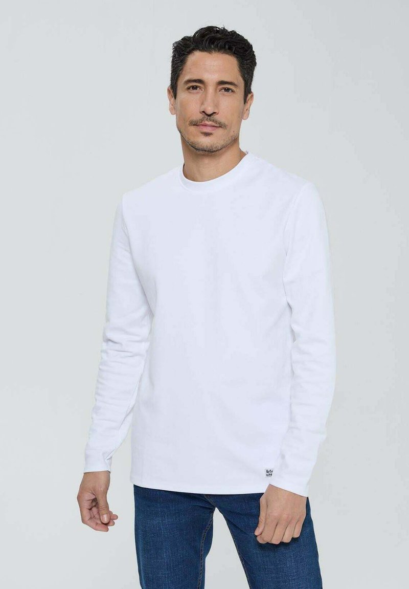 Chemise blanche à manches longues en coton, avec un col rond et une petite étiquette de marque à l'ourlet. Porte avec un jean bleu.