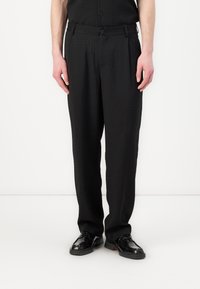 Pantalons noirs avec un motif texturé, coupe droite, plis sur le devant et une taille avec passants de ceinture, associés à des chaussures noires.