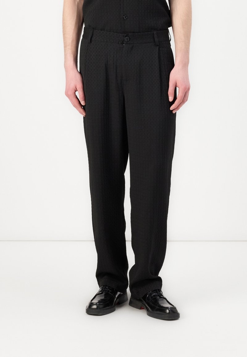 Pantalons noirs avec un motif texturé, coupe droite, plis sur le devant et une taille avec passants de ceinture, associés à des chaussures noires.