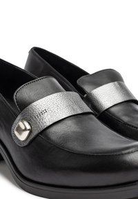 Mocasines de cuero negro con una correa texturizada plateada y acentos metálicos. Acabado suave con punta redonda y suela plana de goma.