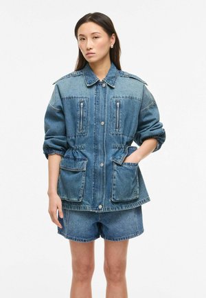 Naine, kes kannab üle suurusega teksajope suurete esitaskutega ja sobivaid teksapükse, seisab lihtsa tausta ees.