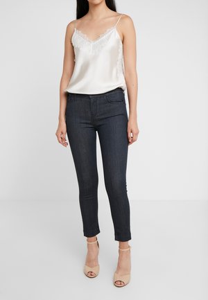 Femme portant un caraco en satin blanc orné de dentelle, un jean skinny foncé et des talons beige à bride cheville et bouts ouverts, debout sur un fond uni.