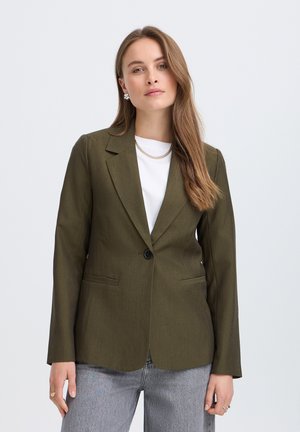 Femme portant un blazer vert olive à un bouton sur un top blanc et un jean gris clair, posant devant un fond clair uni.