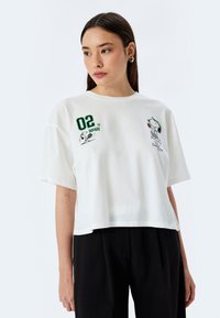 Camiseta de algodón blanca recortada con gráficos de Snoopy en verde y negro, gran texto "02" y mangas cortas holgadas.