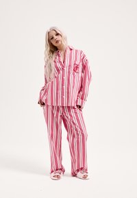 Roza in bela pižama s črtami, ki vključuje srajco z gumbi in žepom, udobne široke hlače ter mehke, puhasto navdahnjene copate.