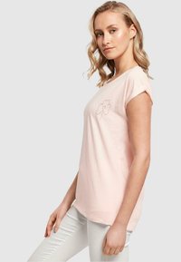 Lys rosa t-skjorte med kort erm som har en enkel svart omriss av en blomst med et hjerte i midten, laget av myk bomullsstoff.