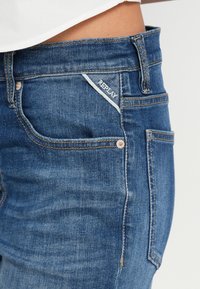 Τα σκούρα μπλε τζιν έχουν εφαρμοστή σχεδίαση, αντίθετη ραφή, μικρή λευκή λεπτομέρεια λογότυπου στην μπροστινή τσέπη και κλασικό στιλ με πέντε τσέπες.