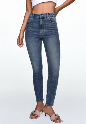 Woman wearing high-waist blue skinny jeans paired with beige heeled sandals and a cropped white top, shown from midriff to feet.