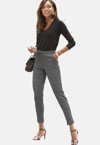 Schwarzer V-Ausschnitt-Pullover kombiniert mit enganliegender Hose im Hahnentritt-Muster. Modell trägt silbernen Schmuck und schwarze Riemchen-High Heels und hält ein Handy.