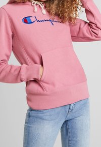 Hoodie rose clair avec un logo brodé bleu et rouge, doté d'une poche avant et d'une capuche avec cordon de serrage. Le tissu semble doux avec des poignets côtelés.