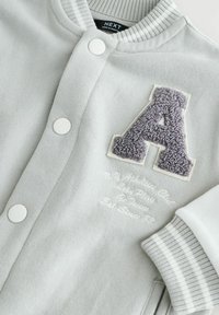 Sweatshirt gris avec des boutons blancs, col et poignets côtelés. Présente un "A" en peluche gris et un texte brodé en blanc en dessous.