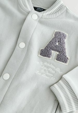 Grå sweatshirt med hvide knapper, ribbestrikket krave og manchetter. Har et luksuriøst gråt "A" og broderet tekst i hvidt nedenfor.