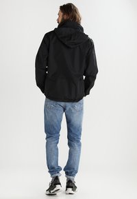 Veste à capuche noire avec des poignets élastiques, associée à un jean bleu clair et des baskets noires. La veste présente une finition lisse et mate.