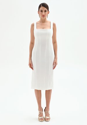 Robe midi blanche moulante avec un décolleté carré, des bretelles larges et des détails de coutures verticales. Associée à des talons dorés à brides.