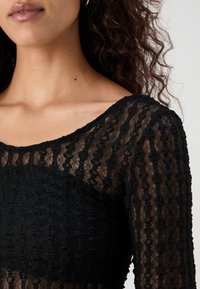 Svart blonde topp med lange ermer og rund hals. Stoffet har teksturerte mønstre og semi-transparente seksjoner.