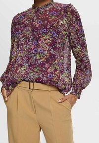 Kwiatowa bluzka na fioletowym tle, z zielonymi, żółtymi i różowymi kwiatami, długimi rękawami oraz falbanami przy dekolcie.