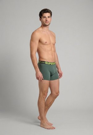 Homme torse nu, debout pieds nus, portant un boxer vert avec une ceinture jaune portant le logo "Superdry" sur un fond gris uni.