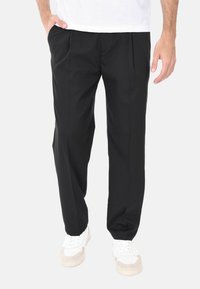 Pantaloni neri a pieghe con vestibilità rilassata, realizzati in un tessuto morbido, abbinati a sneakers bianche. Adatti per un abbigliamento casual o semi-formale.