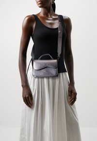 Bolso cruzado de cuero gris con una solapa con patrón de olas, manija superior y correa ajustable, llevado sobre un top negro y una falda blanca.