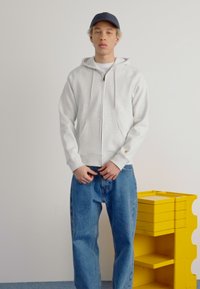 Hellgrauer Reißverschluss-Hoodie mit Kängurutaschen, kombiniert mit lockeren blauen Jeans. Eine marineblaue Cap wird getragen. Gelbe Aufbewahrungseinheit im Hintergrund.