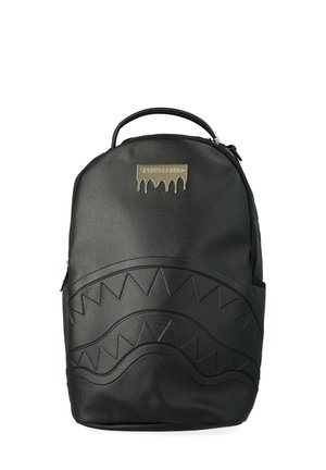 Zaino nero con manico superiore, chiusura con cerniera, motivo a zigzag in rilievo che assomiglia a denti di squalo e una targhetta logo metallica dorata nella parte alta centrale.