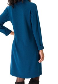 Abito in maglia color teal con colletto alto e polsini a coste. Il design presenta una silhouette rilassata e un orlo fino al ginocchio.