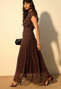 Abito lungo marrone a pois con maniche corte con volant, vita increspata e orlo trasparente. Abbinato a sandali con tacco alto color borgogna e clutch nera.