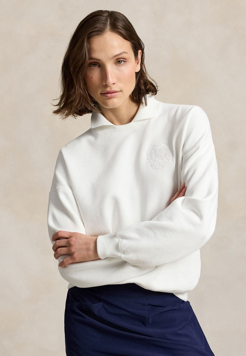 Jeune femme aux cheveux bruns portant un sweat blanc et une jupe bleu marine, les bras croisés, fond neutre.