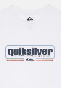 Bílá tričko s logem Quiksilver, modrými a červenými pruhy a textem "ORIGINÁLNÍ SPOLEČNOST NA DESKOVÉ JEŽDĚNÍ" na přední straně.
