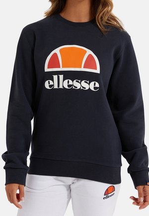 Person trägt einen schwarzen ellesse Sweatshirt mit einem orange-roten Logo und weiße ellesse Hose mit passendem Logo am oberen linken Oberschenkel.